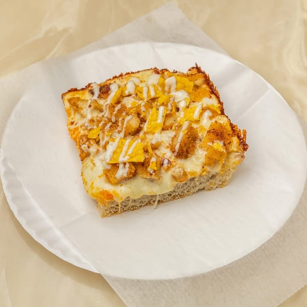 A square slice of Tori T's Pizzeria pizza with melted cheese, chicken, and tortilla chips, drizzled with white sauce.