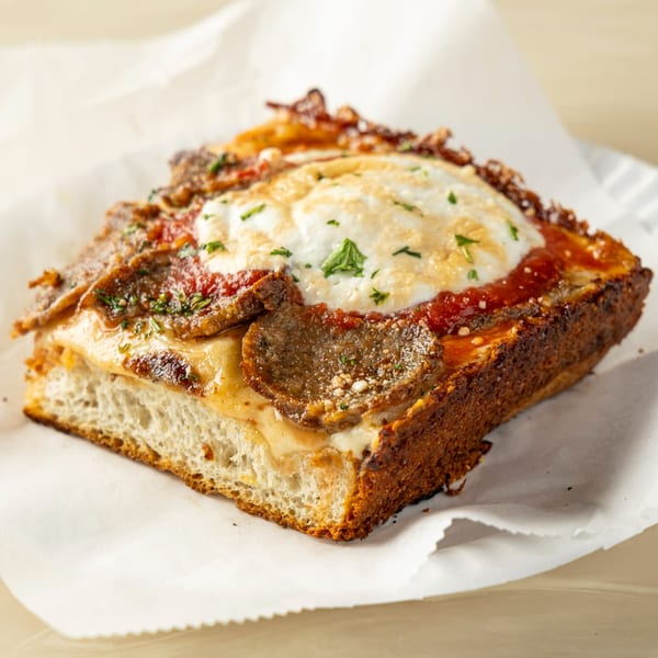 Thick-crust Detroit-style pizza slice with sausage, melted cheese, and red sauce on parchment paper.