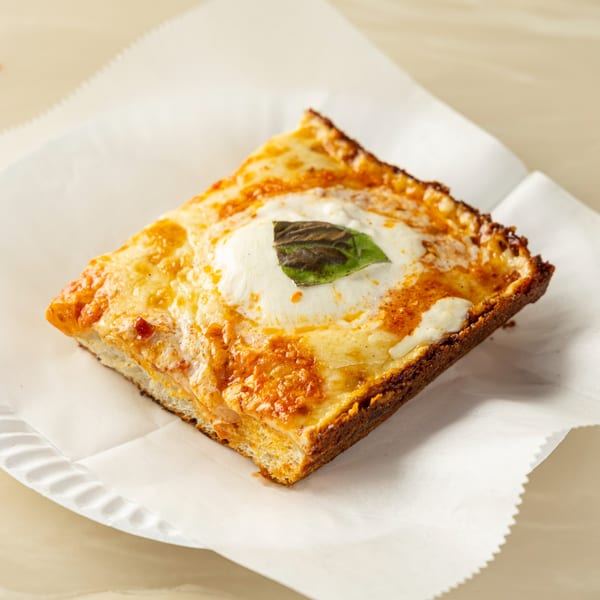Square slice of pizza with melted cheese, tomato sauce, and a basil leaf on parchment paper.